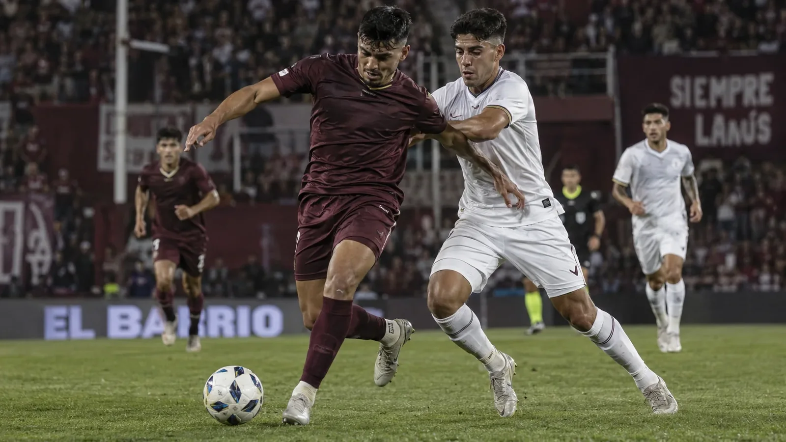Partido de fútbol nocturno en un estadio, con equipos de camisetas granate y blancas sin escudos identificables; imagen ilustrativa.